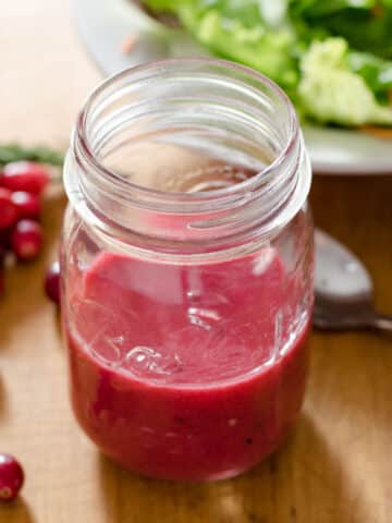 Cranberry salad dressing in mason jar next to cranberries, spoon and lettuce.