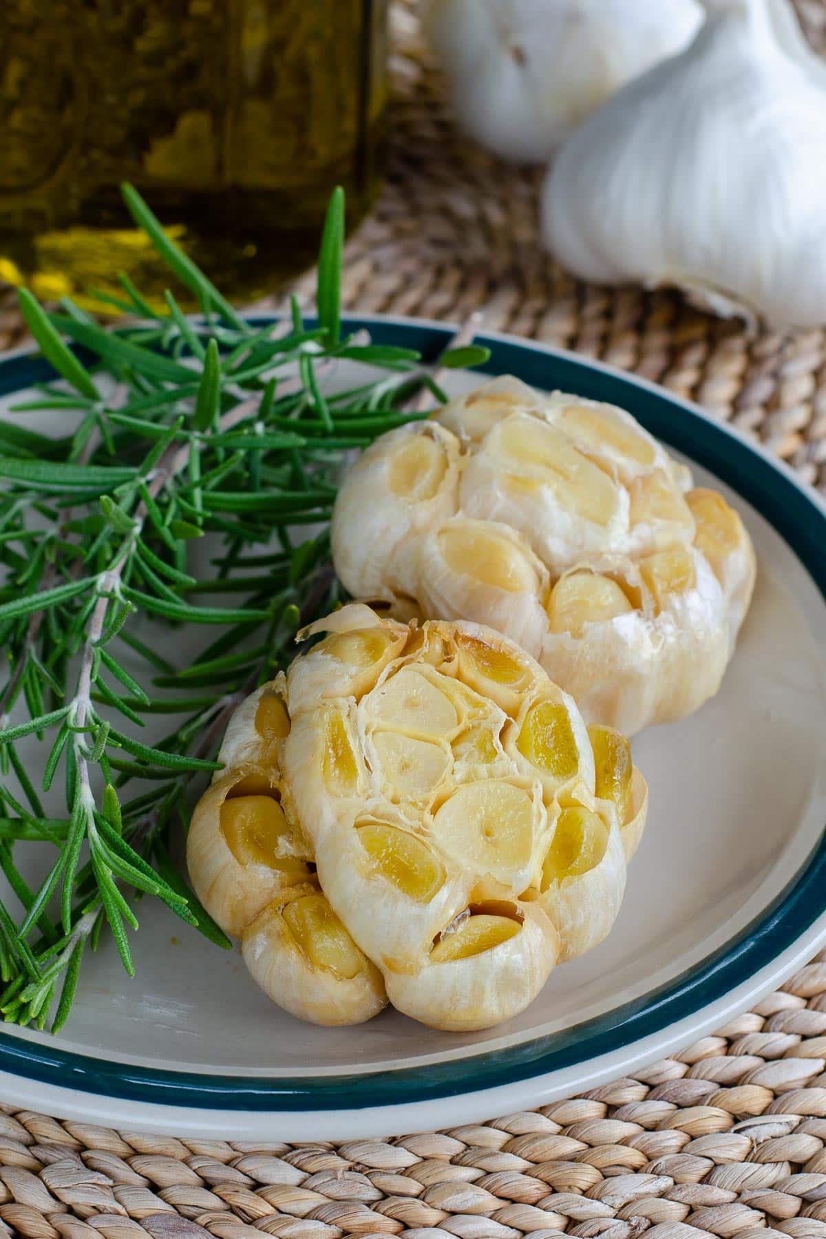 Roasted garlic heads on plate with rosemary, olive oil and whole bulbs in background
