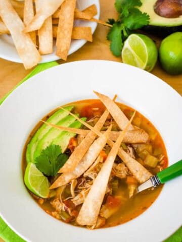 Bowl of chicken tortilla soup with crispy tortilla strips, limes, avocado and cilantro