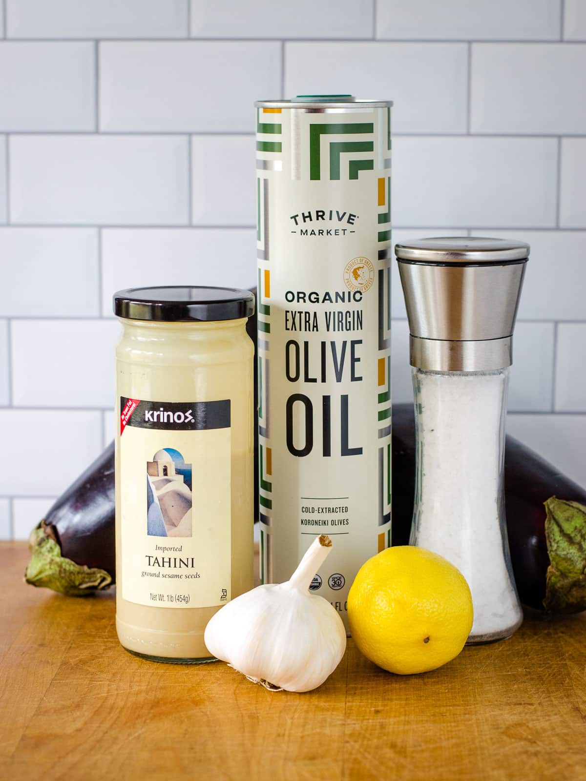 Jar of tahini, can of olive oil, salt shaker, head of garlic, one lemon and two eggplants on cutting board.