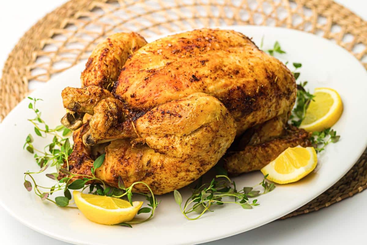 Roast chicken on a white platter with herbs and lemon wedges.