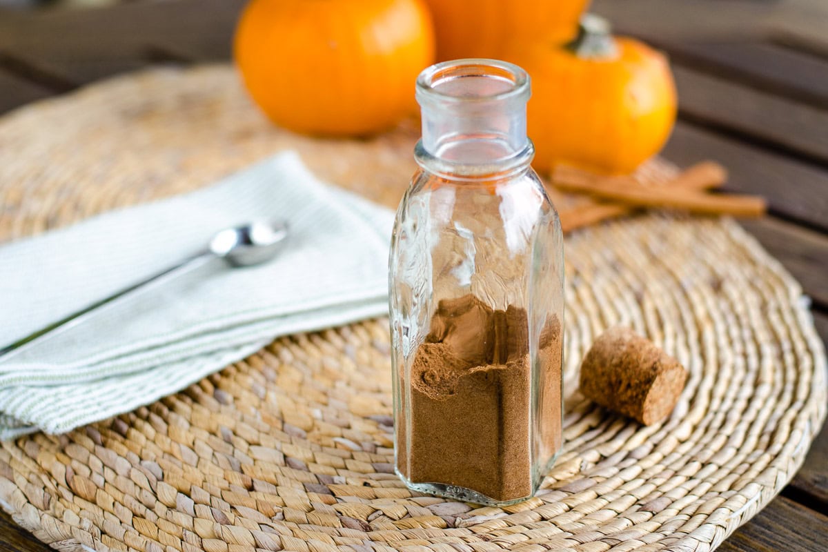 Homemade pumpkin pie spice in glass bottle with mini pumpkins, cinnamon sticks, teaspoon and cork on woven placemat.