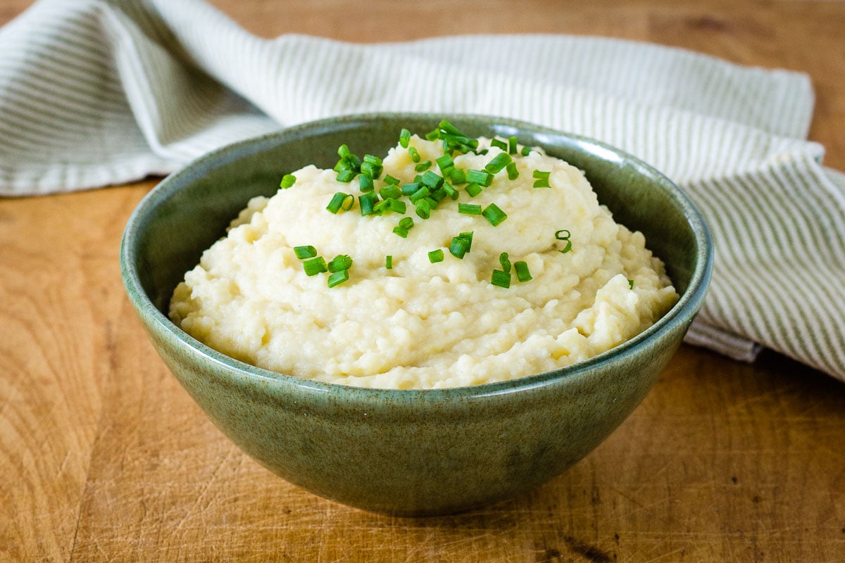 Slow cooker mashed potatoes garnished with chives in green bowl on board with napkin in background.