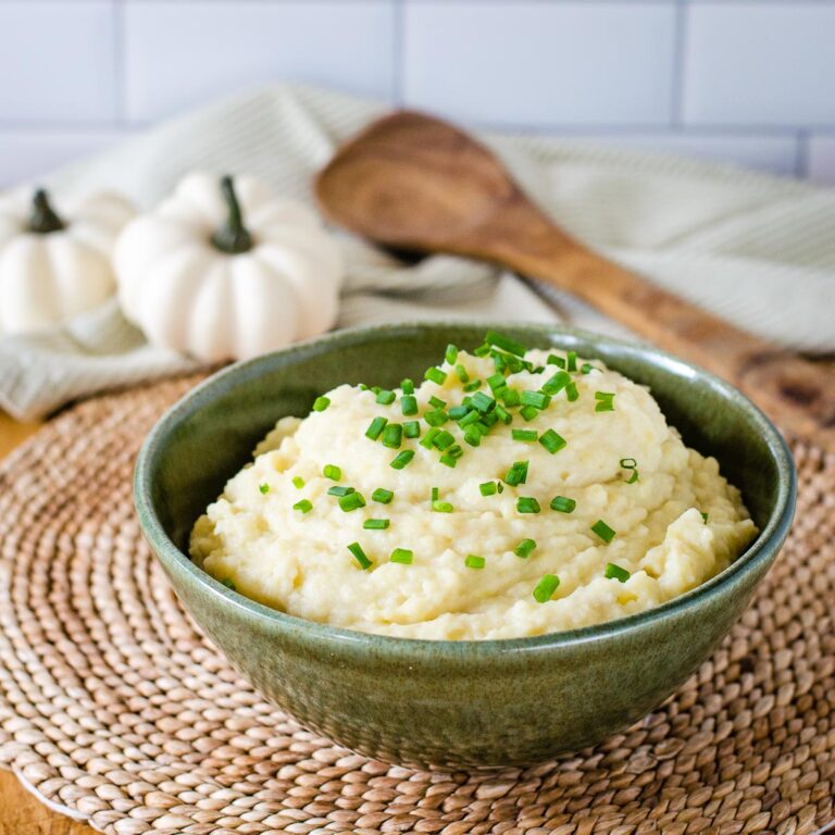 Slow cooker mashed potatoes garnished with chives in green bowl with white pumpkins in background.