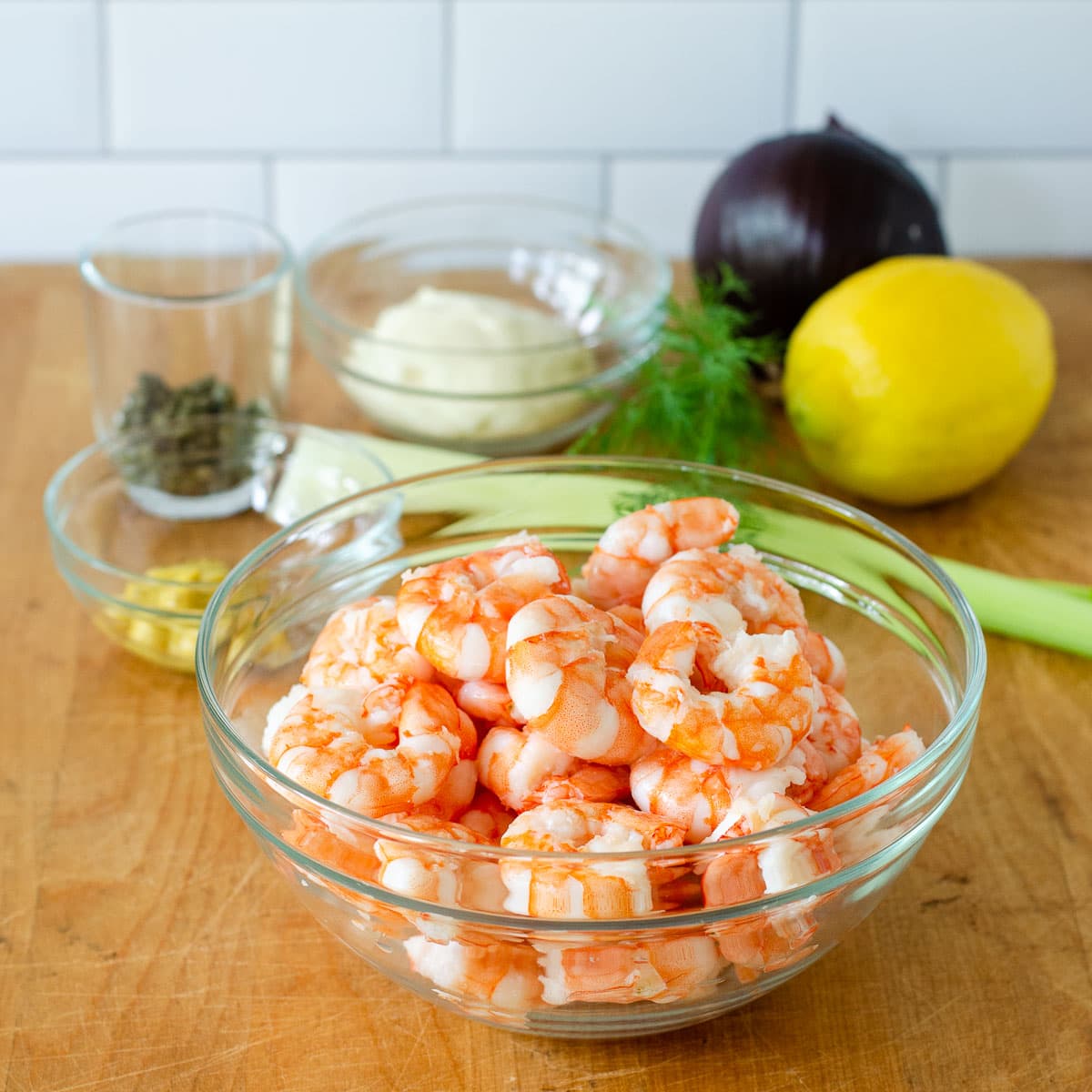 Bowls of cooked shrimp, mustard, capers, and mayonnaise, next to a purple onion, lemon, fresh dill and celery ribs.