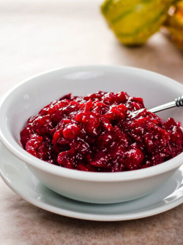 Cranberry sauce in white bowl with saucer and spoon, and cranberries in the background.