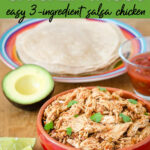 Bowl of shredded crockpot salsa chicken topped with fresh cilantro, with a stack of tortillas, avocado, lime wedges and salsa on a wooden board for chicken tacos. Text is Crockpot Chicken Tacos, easy 3-ingredient salsa chicken.