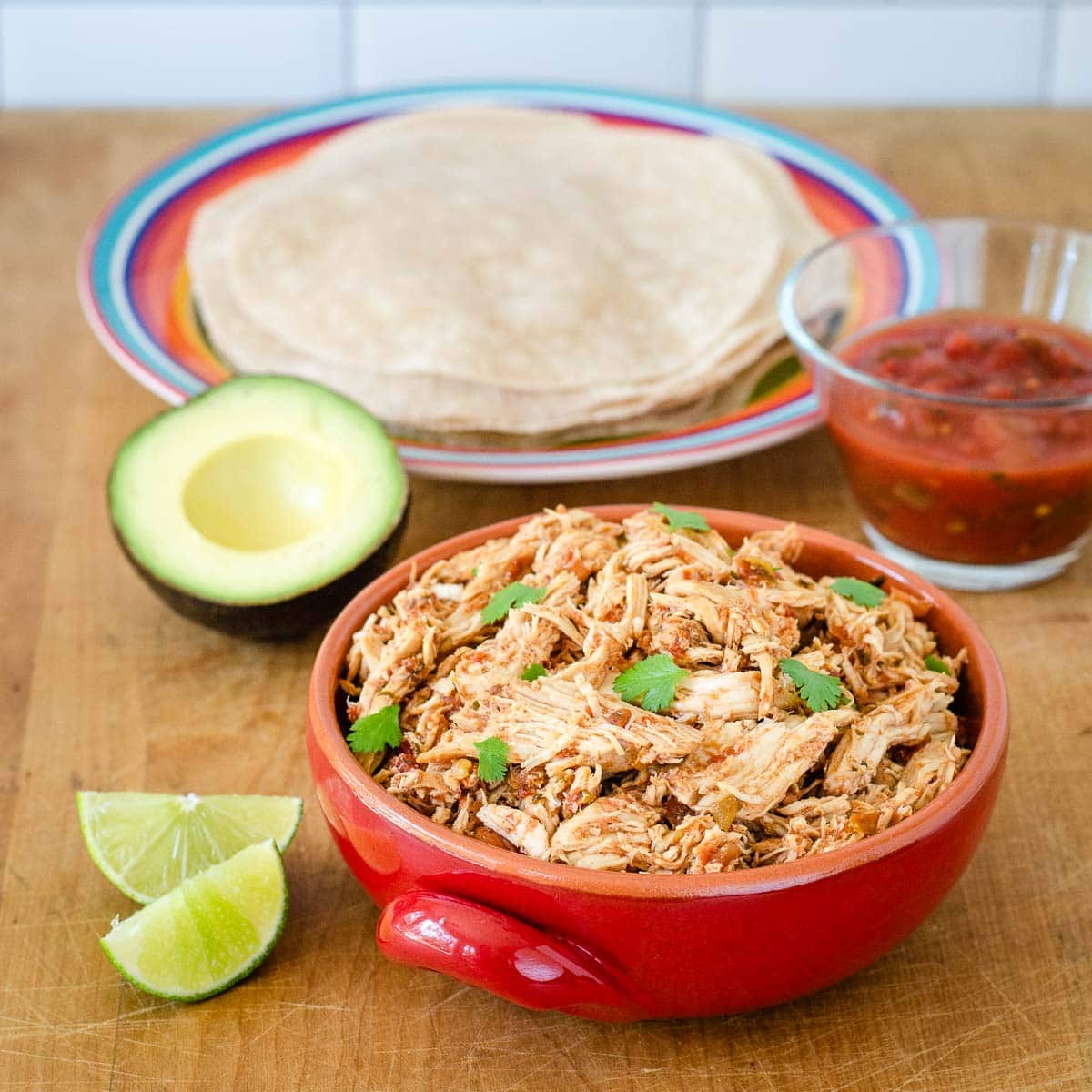 Crockpot salsa chicken in a bowl next to tortillas, salsa, avocado and lime.