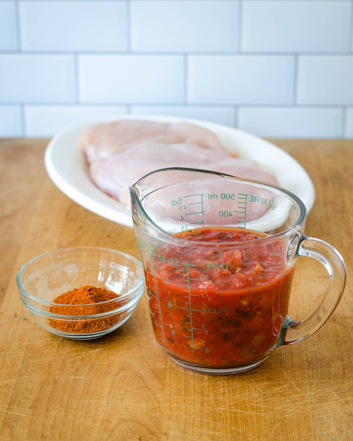 Measuring cup with salsa, small bowl with chili powder and three raw boneless, skinless chicken breasts on a plate.