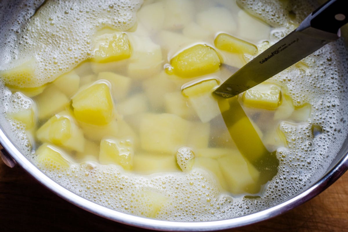 Pot of boiled potato cubes with knife testing doneness.